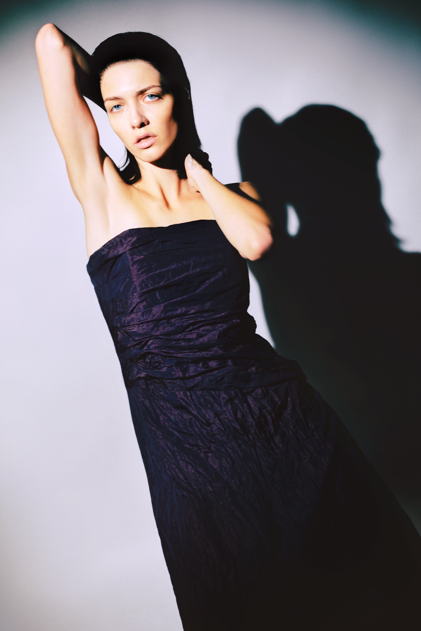 Portret kobiety w fioletowej sukni bez ramiączek i kapeluszu, pozującej na jasnym tle z wyraźnym cieniem. Ujęcie studyjne z mocnym światłem podkreślającym rysy twarzy.