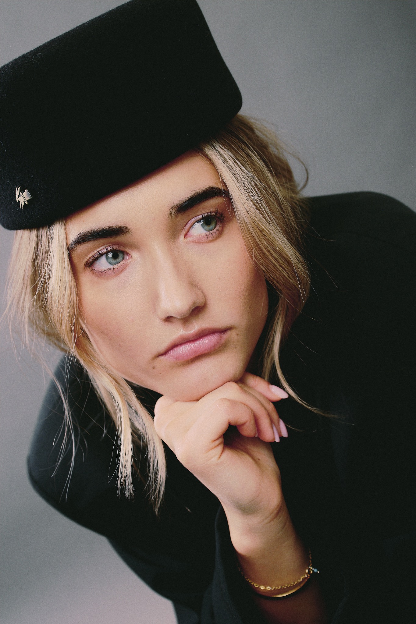 Portret kobiety z blond włosami, w czarnym kapeluszu i golfie, opierającej brodę na dłoni. Delikatny makijaż, jasne oczy, złota bransoletka na nadgarstku.
