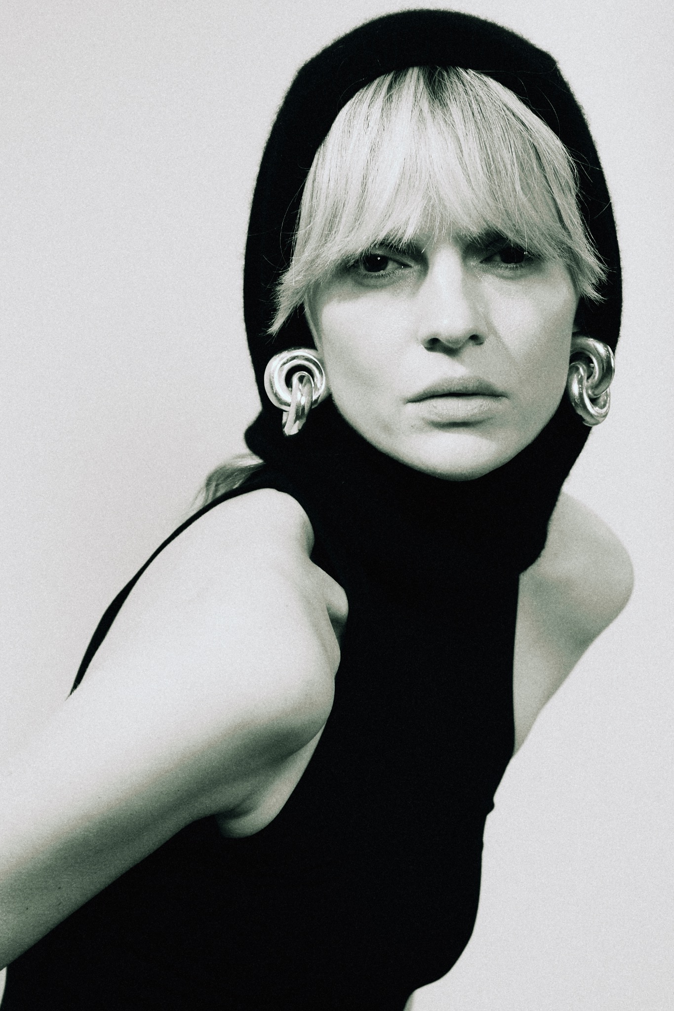 Portret kobiety z blond włosami w czarnym golfie i kapturze, z dużymi srebrnymi kolczykami na białym tle. Ujęcie z bliska, ostre światło.