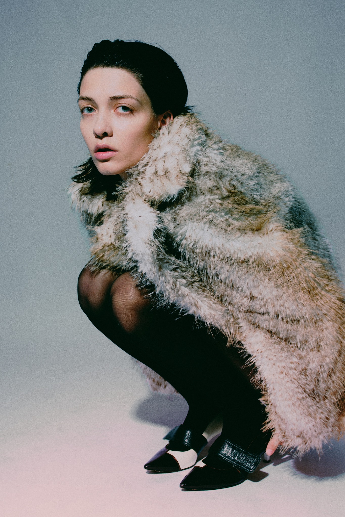 Kobieta w futrze pozuje w kucki, patrząc w obiektyw podczas sesji w Niżnej Łące. Uwagę przykuwają buty z paskiem i jasnymi wstawkami.