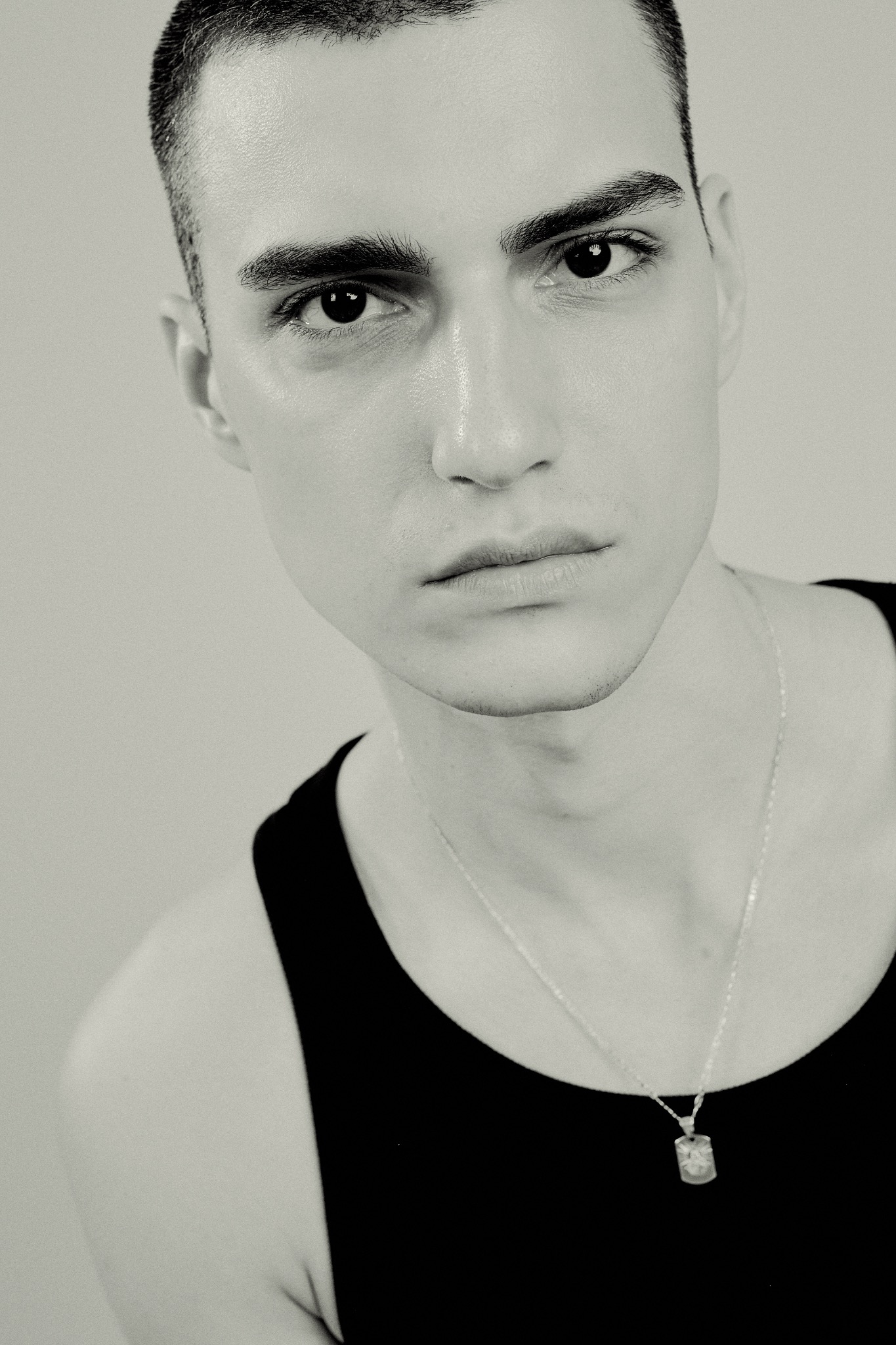 Czarno-biały portret młodego mężczyzny z krótkimi włosami, w czarnym podkoszulku i srebrnym łańcuszku, skierowany w stronę obiektywu, z neutralnym wyrazem twarzy.