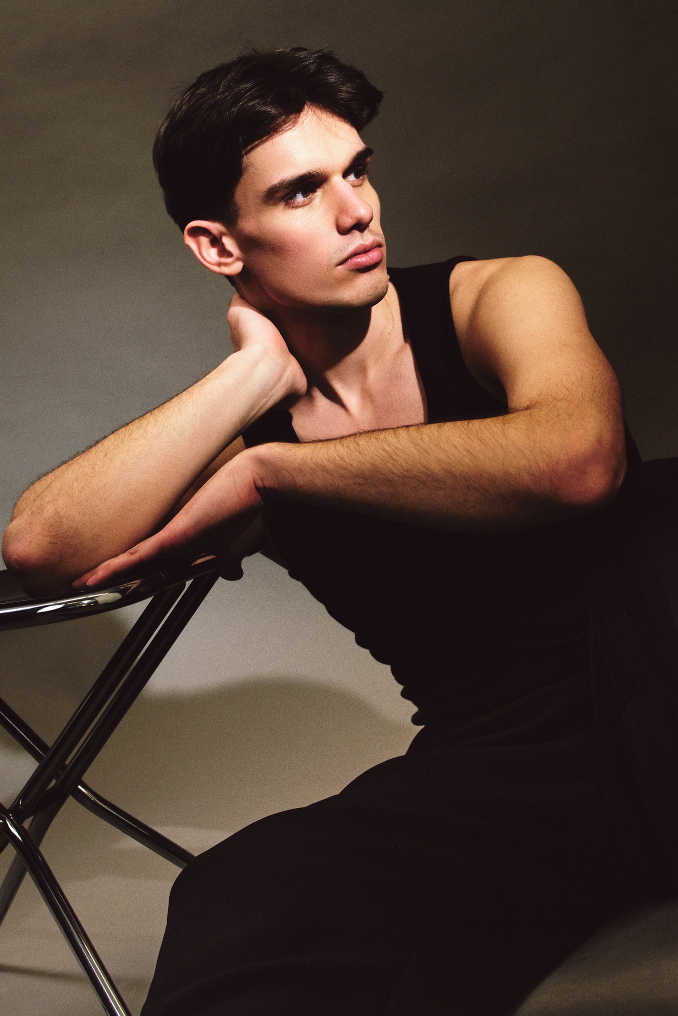 Mężczyzna w czarnym podkoszulku opiera się o metalowe krzesło, spoglądając w bok. Portret w stonowanej kolorystyce, z delikatnym oświetleniem.