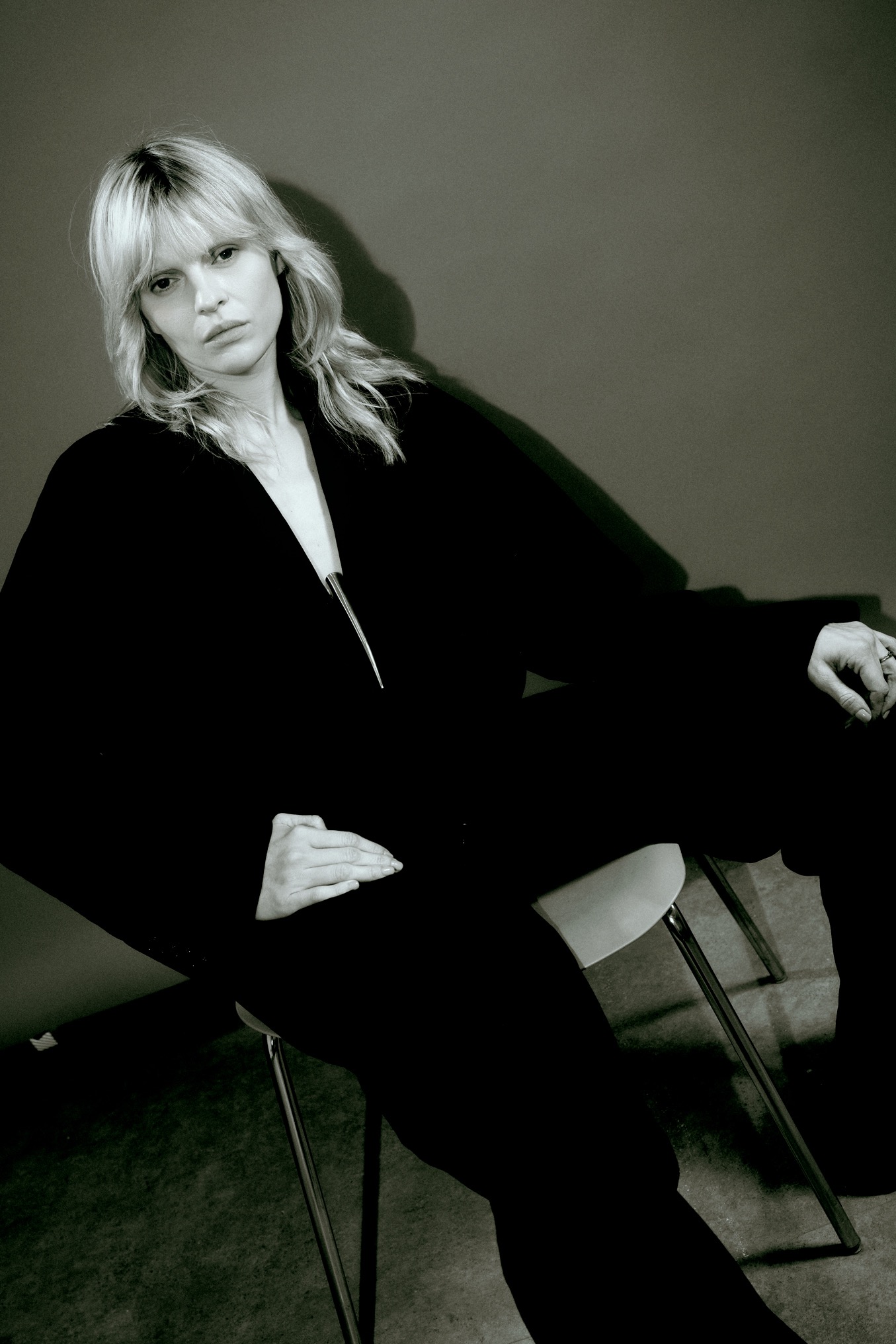 Monochromatyczne ujęcie kobiety w czarnym garniturze, siedzącej na krześle. Minimalistyczne tło, ostre światło podkreśla rysy twarzy i fakturę materiału.