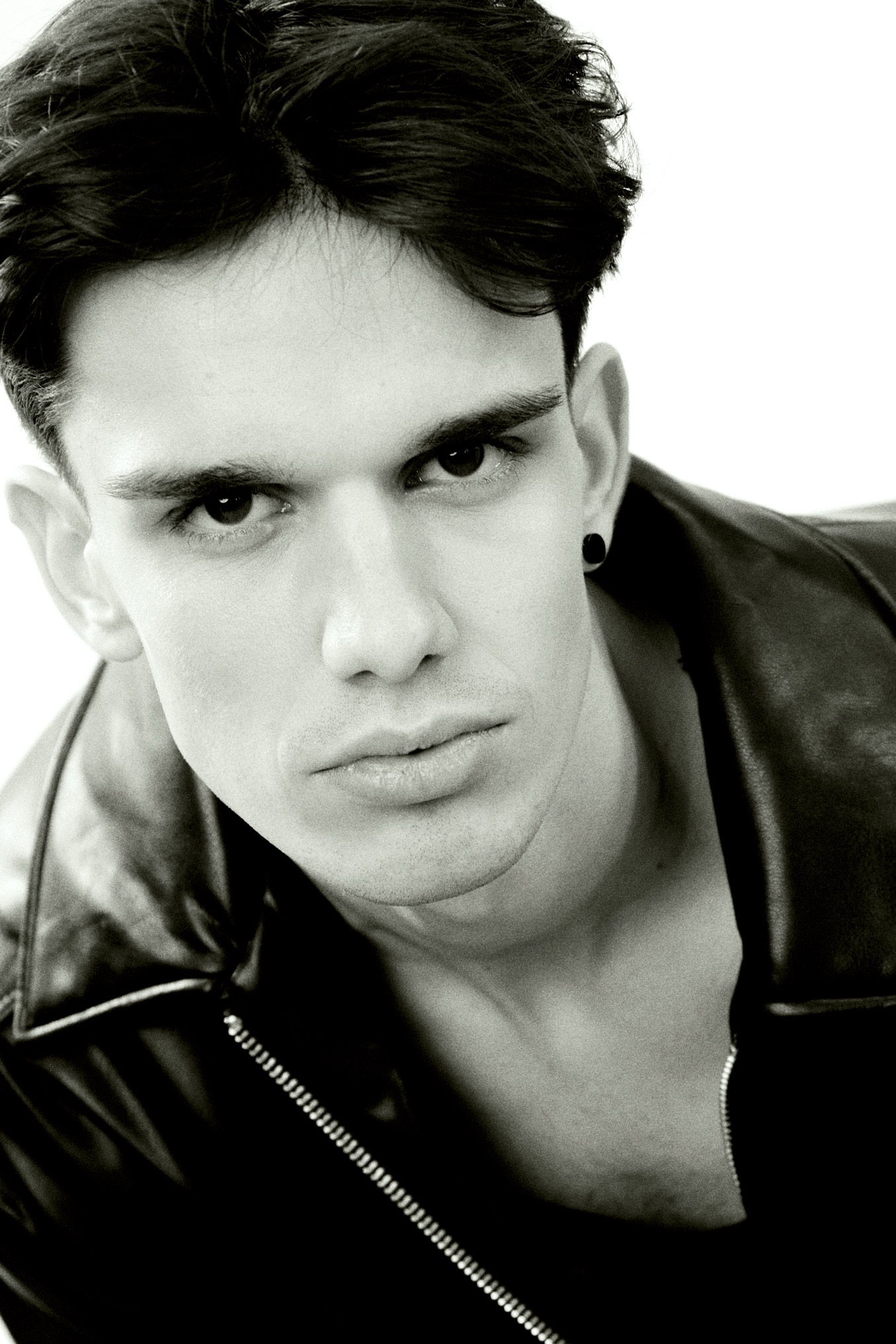 Czarno-biały portret młodego mężczyzny w skórzanej kurtce, z kolczykiem w uchu, spoglądającego w górę, z mocnym oświetleniem i wyraźnymi rysami twarzy.
