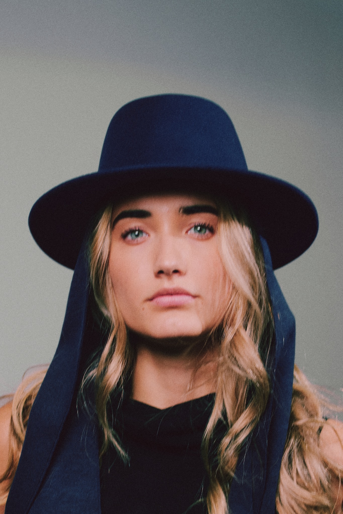 Portret kobiety o jasnych włosach i niebieskich oczach, w ciemnym kapeluszu i szalu, na jasnym tle. Ujęcie z bliska, skupione na twarzy.