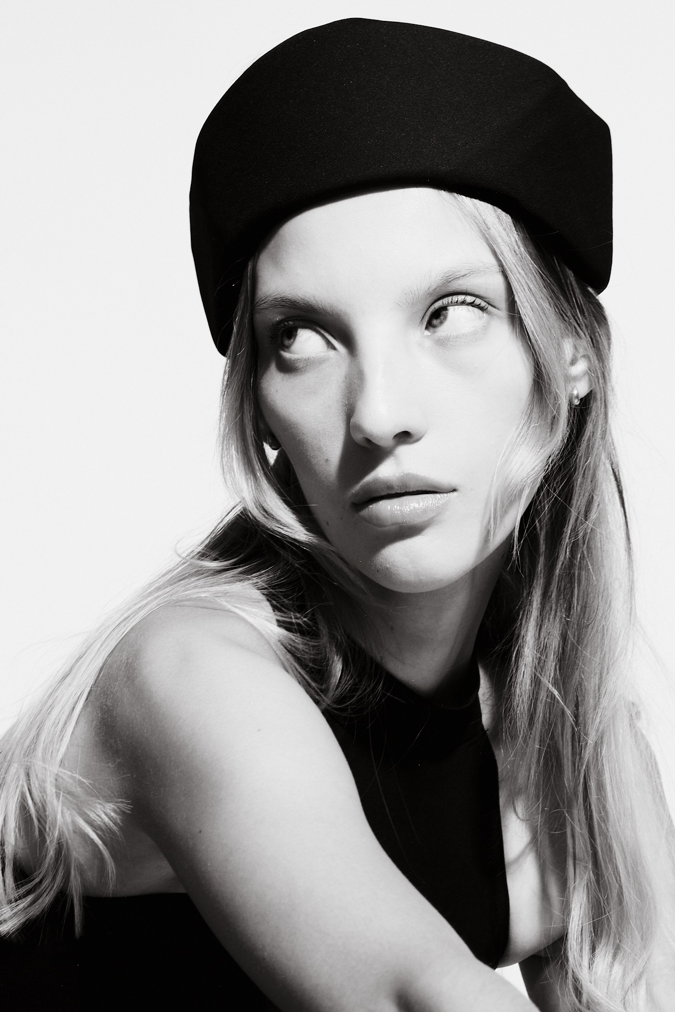 Czarno-biały portret kobiety w czarnym berecie, z długimi blond włosami, spoglądającej w lewo, na jasnym tle. Minimalistyczna estetyka, podkreślone światłocienie.