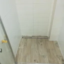 Nowoczesny prysznic walk-in z odpływem liniowym i płytkami imitującymi drewno na podłodze. Białe, strukturalne płytki na ścianach. Minimalistyczne wykończenie.