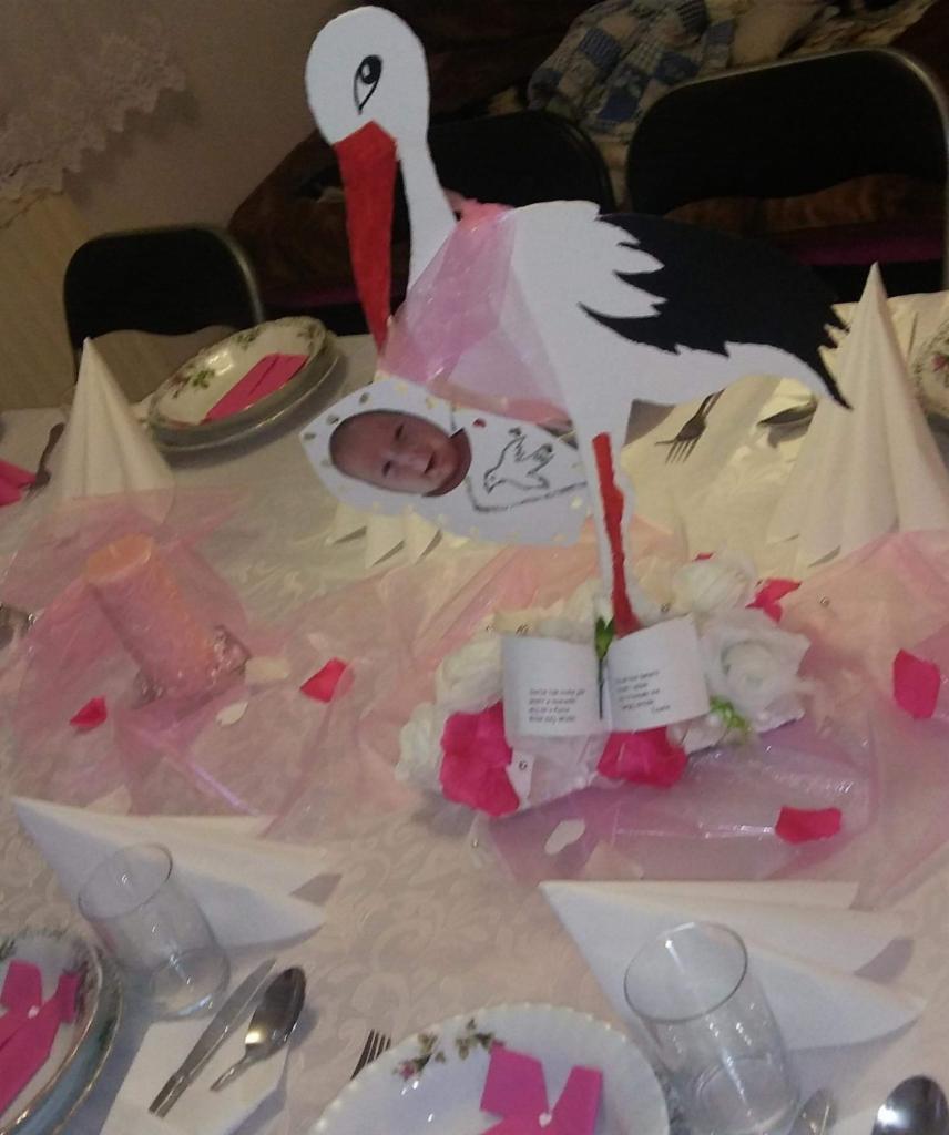 Dekoracja stołu z bocianem trzymającym zdjęcie dziecka, różowe akcenty, białe serwetki, zastawa stołowa i świece.
