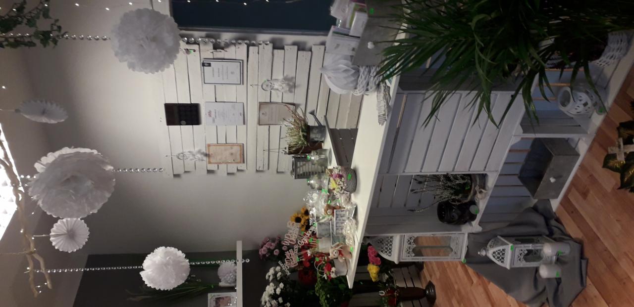 Dekoracje sali: białe pompony zawieszone na srebrnych łańcuszkach, drewniana ściana z certyfikatami, kwiaty i dekoracje na białym regale z drewnianych desek.