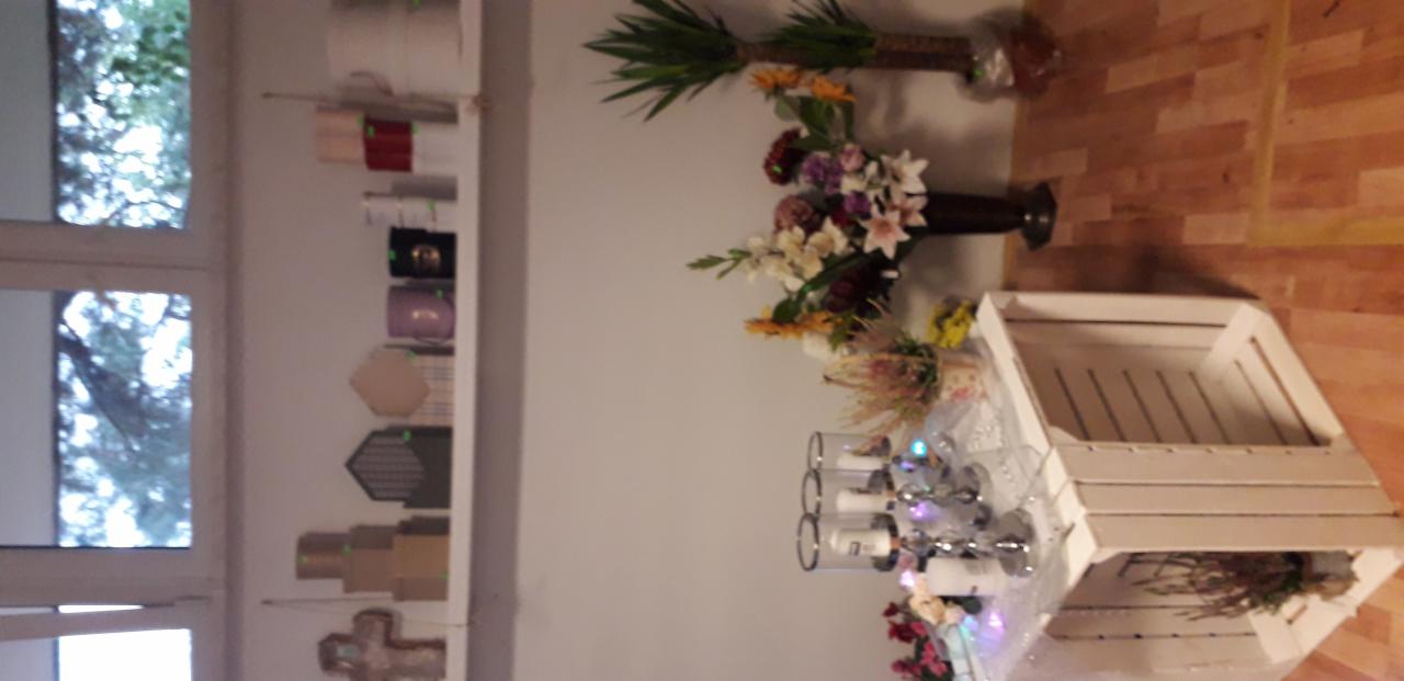 Dekoracja stołu z kwiatami w wazonie, świecznikami i drewnianą skrzynką na tle ściany z półkami z pudełkami i rośliną doniczkową.