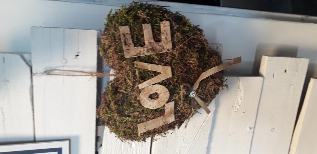 Dekoracja ścienna w kształcie serca z mchu, z napisem LOVE wykonanym z kory brzozy i ozdobną kokardką z kryształkiem, zawieszona na białych drewnianych panelach.