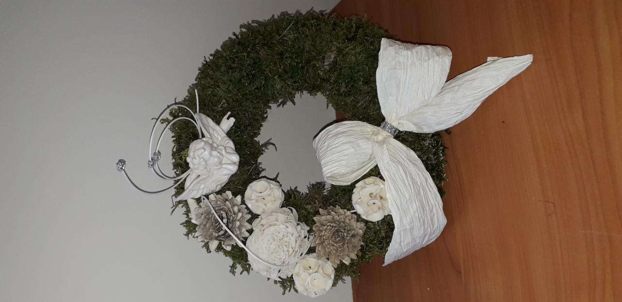Wieniec z mchu, białych kwiatów i aniołka, ozdobiony kremową, papierową kokardą z błyszczącym akcentem.