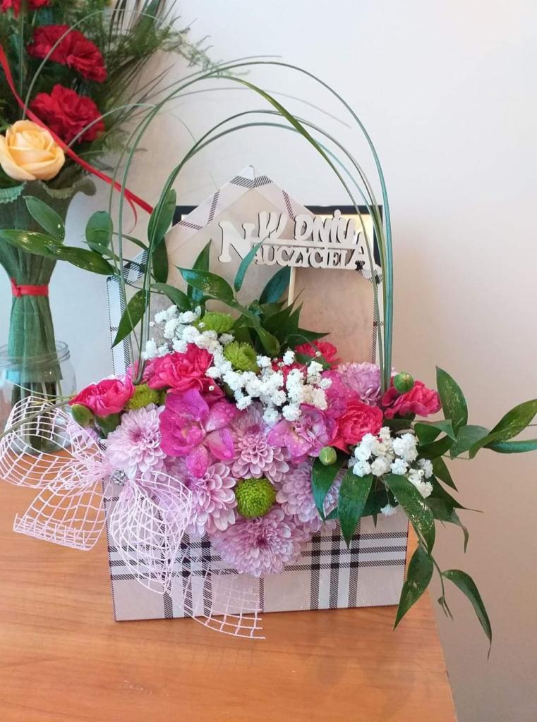 Kompozycja kwiatowa w pudełku w kratę z napisem 'Na Dzień Nauczyciela', z różowymi, białymi i zielonymi kwiatami, przewiązana delikatną wstążką.