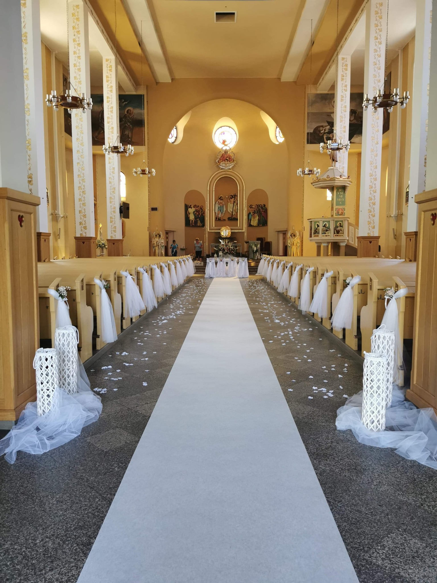 Uroczysty wystrój kościoła przed ceremonią ślubną: biały dywan, białe dekoracje na ławkach, lampiony i rozsypane płatki kwiatów.