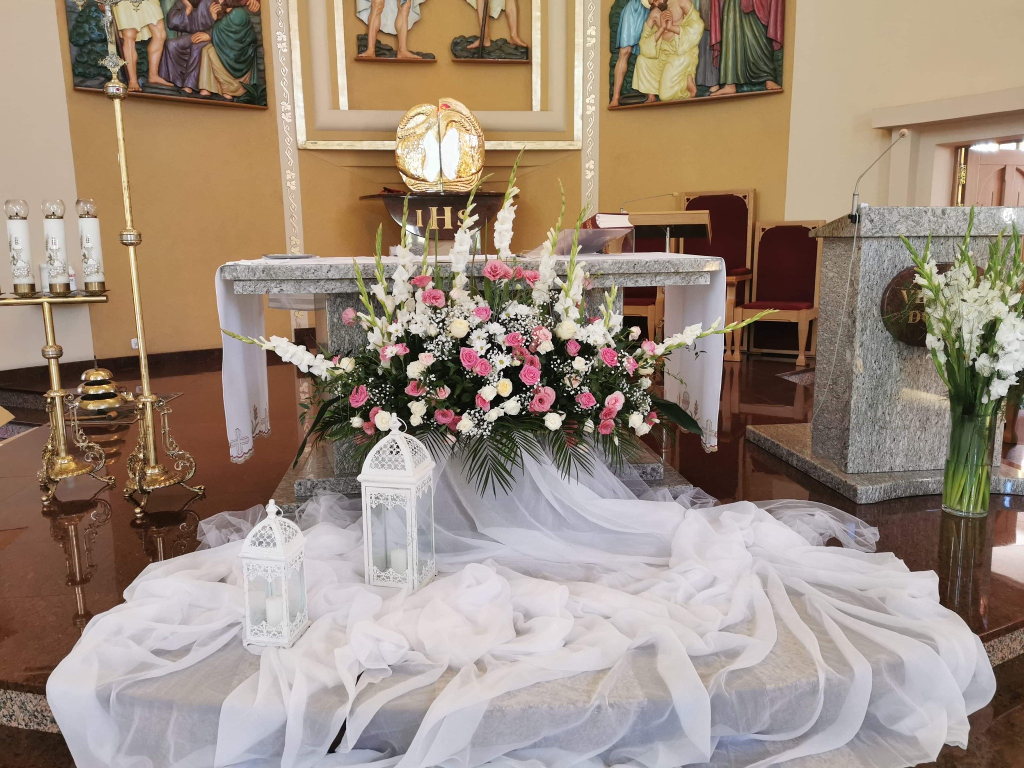 Dekoracja kwiatowa z białymi lampionami i zwiewną tkaniną na ołtarzu w kościele.