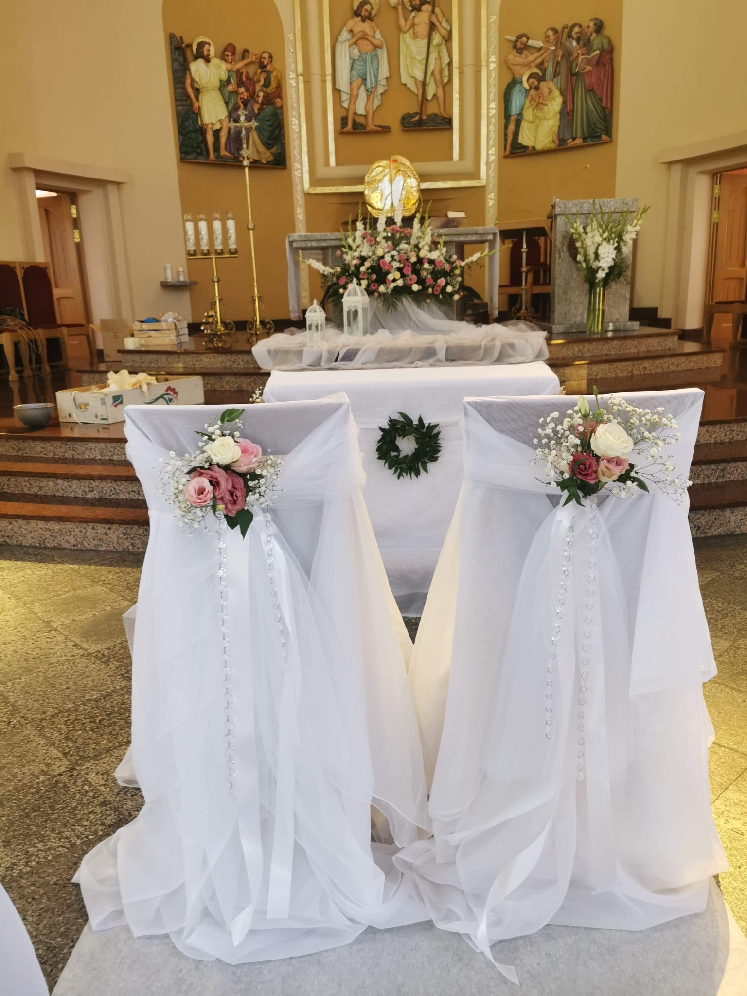 Udekorowane krzesła w kościele na uroczystość ślubną, z białymi pokrowcami, kwiatowymi kompozycjami i girlandami z perełek, w tle ołtarz z kwiatami i obrazami religijnymi.