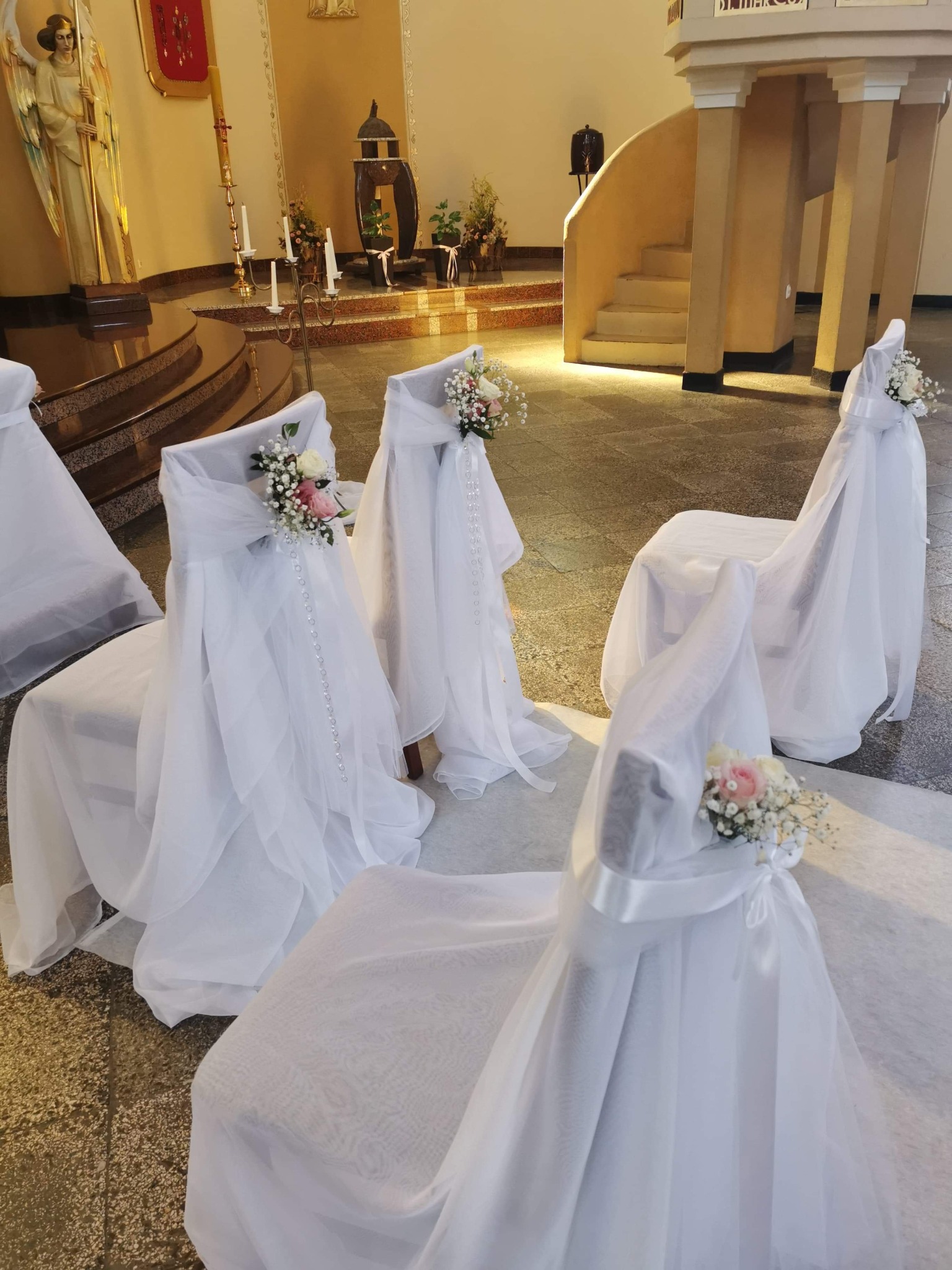 Dekoracje krzeseł w kościele na uroczystość ślubną, białe pokrowce z tiulu, kwiatowe kompozycje z różowych i białych róż oraz drobnych kwiatów, perłowe girlandy.