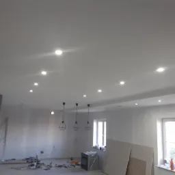 Białe wnętrze w trakcie prac wykończeniowych z widocznymi przewodami, narzędziami i płytami gipsowymi na podłodze, oświetlone punktowymi lampami w suficie i trzema lampami wiszącymi...