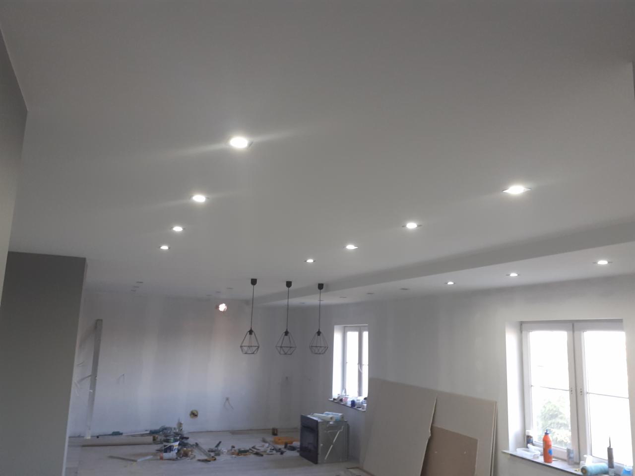 Białe wnętrze w trakcie prac wykończeniowych z widocznymi przewodami, narzędziami i płytami gipsowymi na podłodze, oświetlone punktowymi lampami w suficie i trzema lampami wiszącymi...