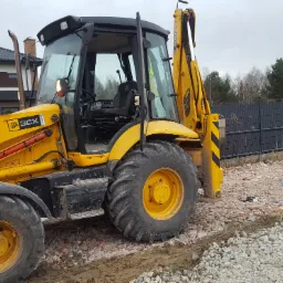 Żółta koparko-ładowarka JCB 3CX na terenie budowy, widoczny fragment budynku mieszkalnego w tle, teren przygotowany pod budowę, wysypany gruzem i kamieniami.