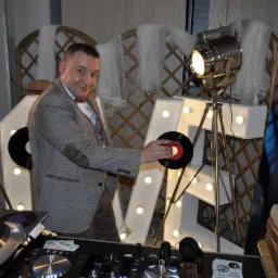 Uśmiechnięty mężczyzna w tweedowej marynarce z łatami na łokciach, trzymający winylową płytę na tle dekoracyjnych, podświetlanych liter i reflektorów studyjnych, z widocznym sprzętem DJ-skim.