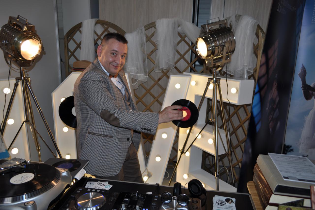 Uśmiechnięty mężczyzna w tweedowej marynarce z łatami na łokciach, trzymający winylową płytę na tle dekoracyjnych, podświetlanych liter i reflektorów studyjnych, z widocznym sprzętem DJ-skim.