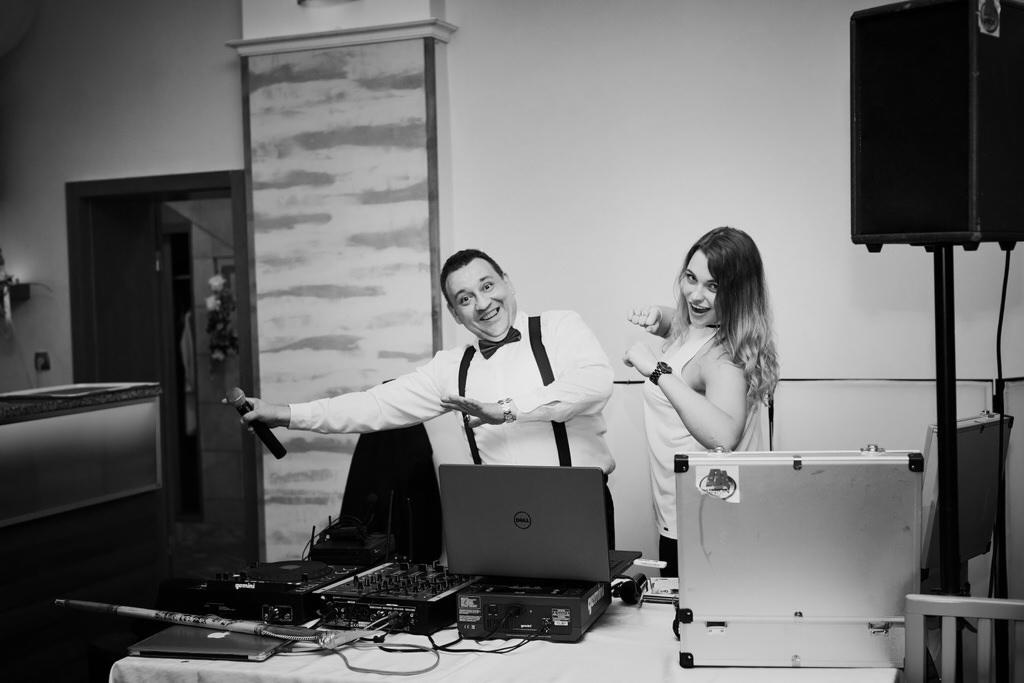 Mężczyzna z mikrofonem i kobieta w tle, sprzęt DJ-ski, laptop Dell, kolumna głośnikowa na statywie, czarno-białe zdjęcie.