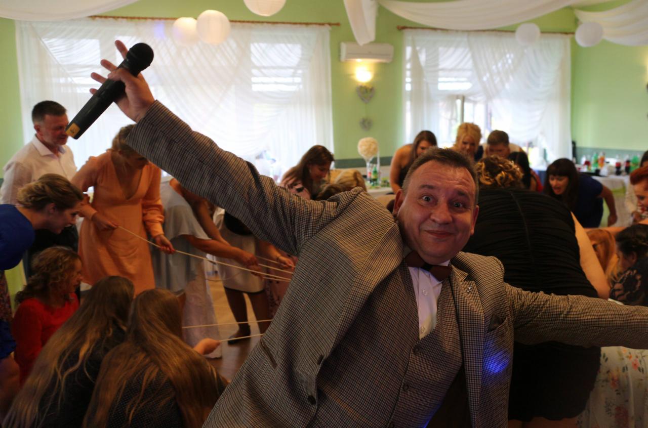 Uśmiechnięty mężczyzna w tweedowej marynarce z muszką, trzymający mikrofon nad głową, prowadzi zabawę z grupą osób na sali weselnej, z białymi lampionami i dekoracjami w tle.