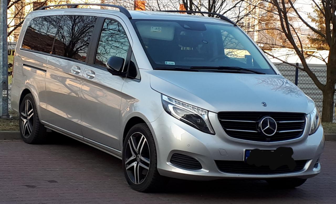 Srebrny van Mercedes-Benz V-klasy zaparkowany na chodniku z kostki, widok z przodu, zarys drzewa odbity w szybach.