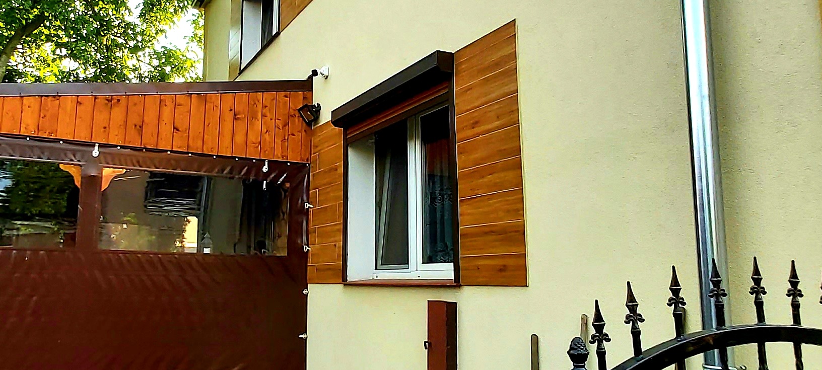 Kamera monitoringu zamontowana na elewacji domu obok drewnianej pergoli z brązową plandeką i okna z drewnianymi okiennicami.