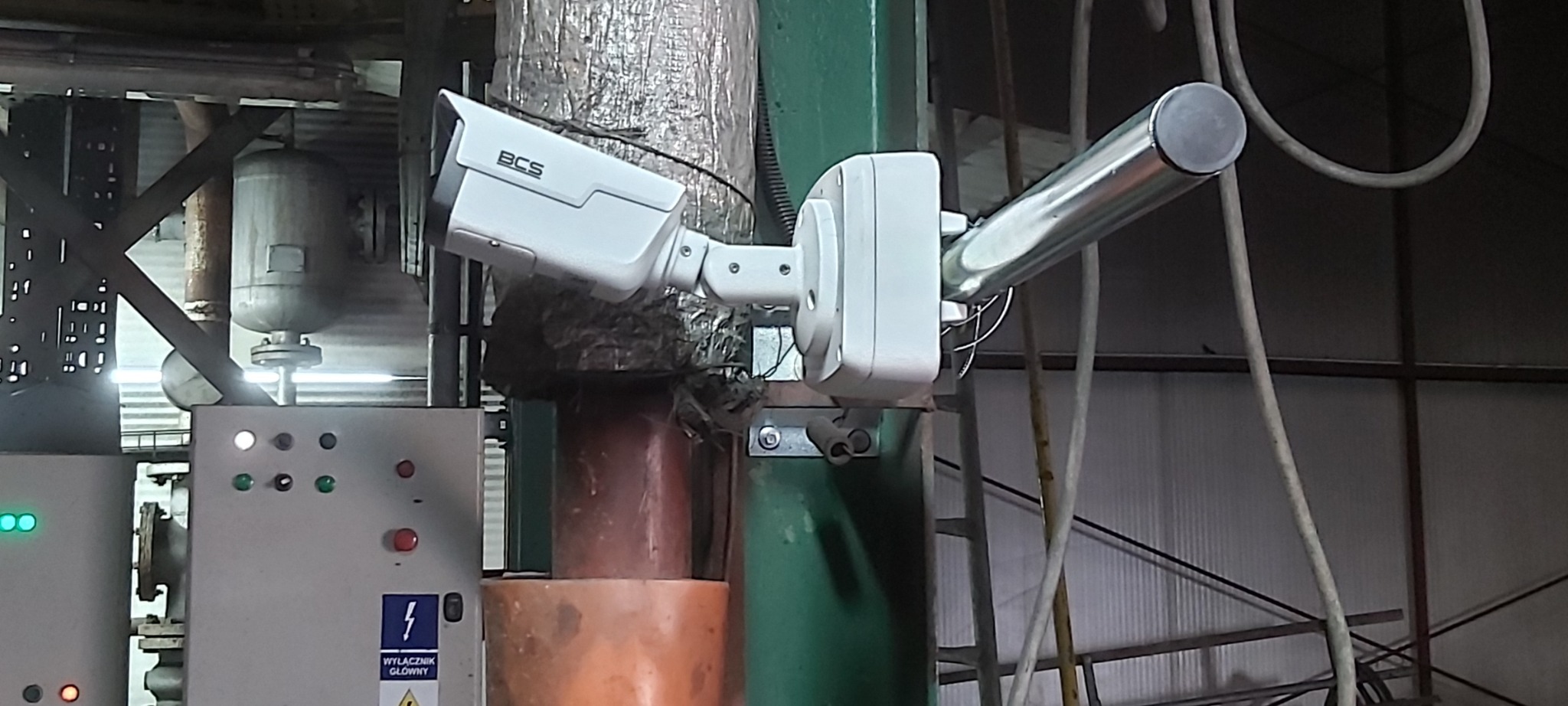 Kamera monitoringu BCS zamontowana w hali przemysłowej, widoczne rury, kable, elementy konstrukcyjne hali i panel sterowania.