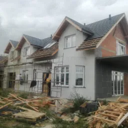 Dom w trakcie prac elewacyjnych: częściowo ocieplony styropianem, z nałożonym szarym tynkiem, widoczne rusztowania, niezabezpieczone drewniane elementy konstrukcji dachu oraz porozrzucane deski...