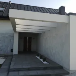 Nowoczesny dom z białą elewacją, ciemnym dachem i zadaszonym wejściem z szarą kostką brukową, widoczne ślady po niedawno zakończonym ocieplaniu elewacji.