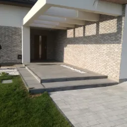 Nowoczesny dom z elewacją z jasnej cegły, zadaszonym wejściem z białymi belkami i schodami wyłożonymi szarymi płytami, otoczony trawnikiem i ścieżką z kostki brukowej.