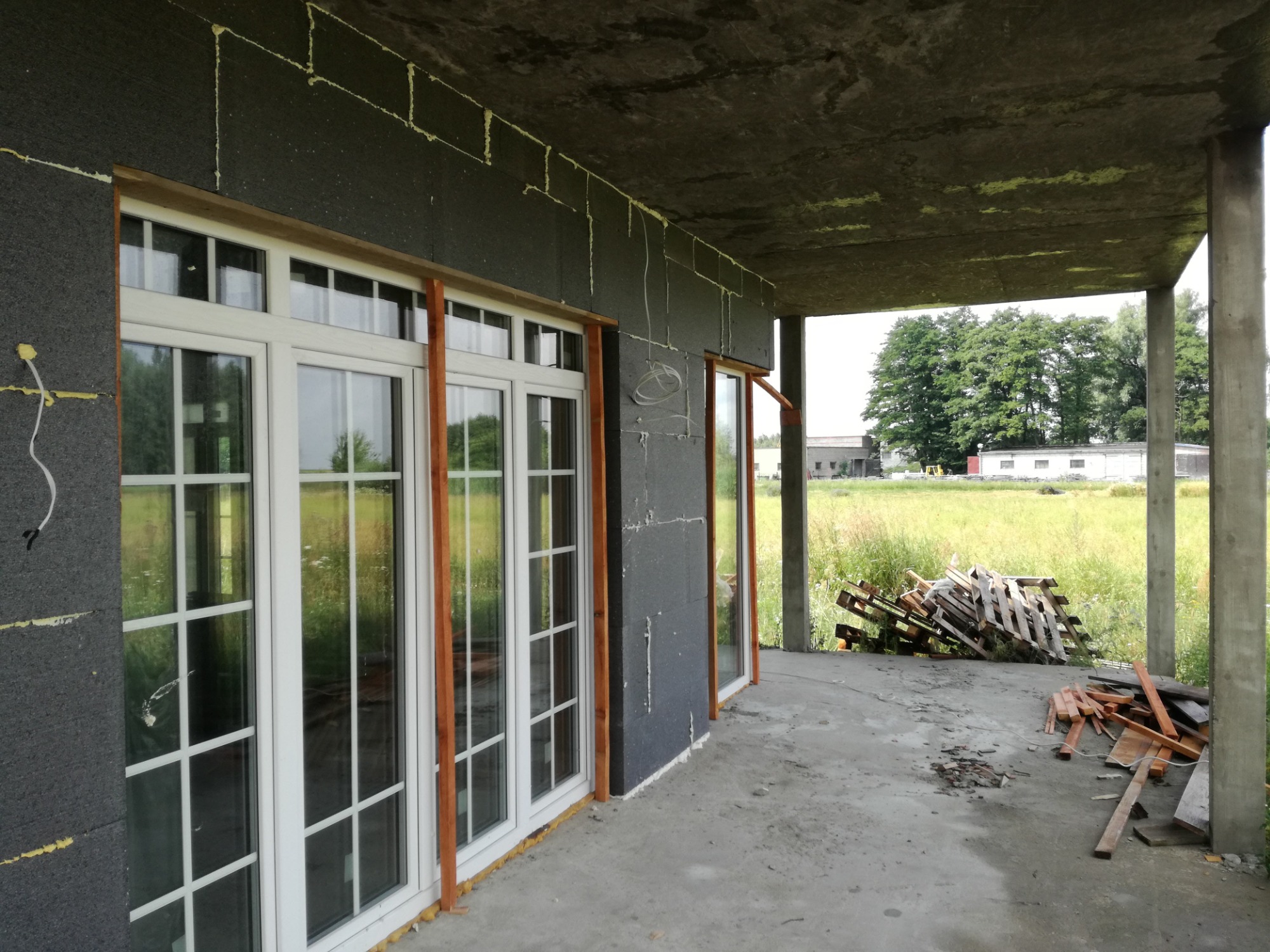 Montaż stolarki okiennej w budynku z częściowo ocieploną elewacją styropianem grafitowym, widoczne drewniane listwy przy oknach i luźno ułożone palety na zewnątrz.