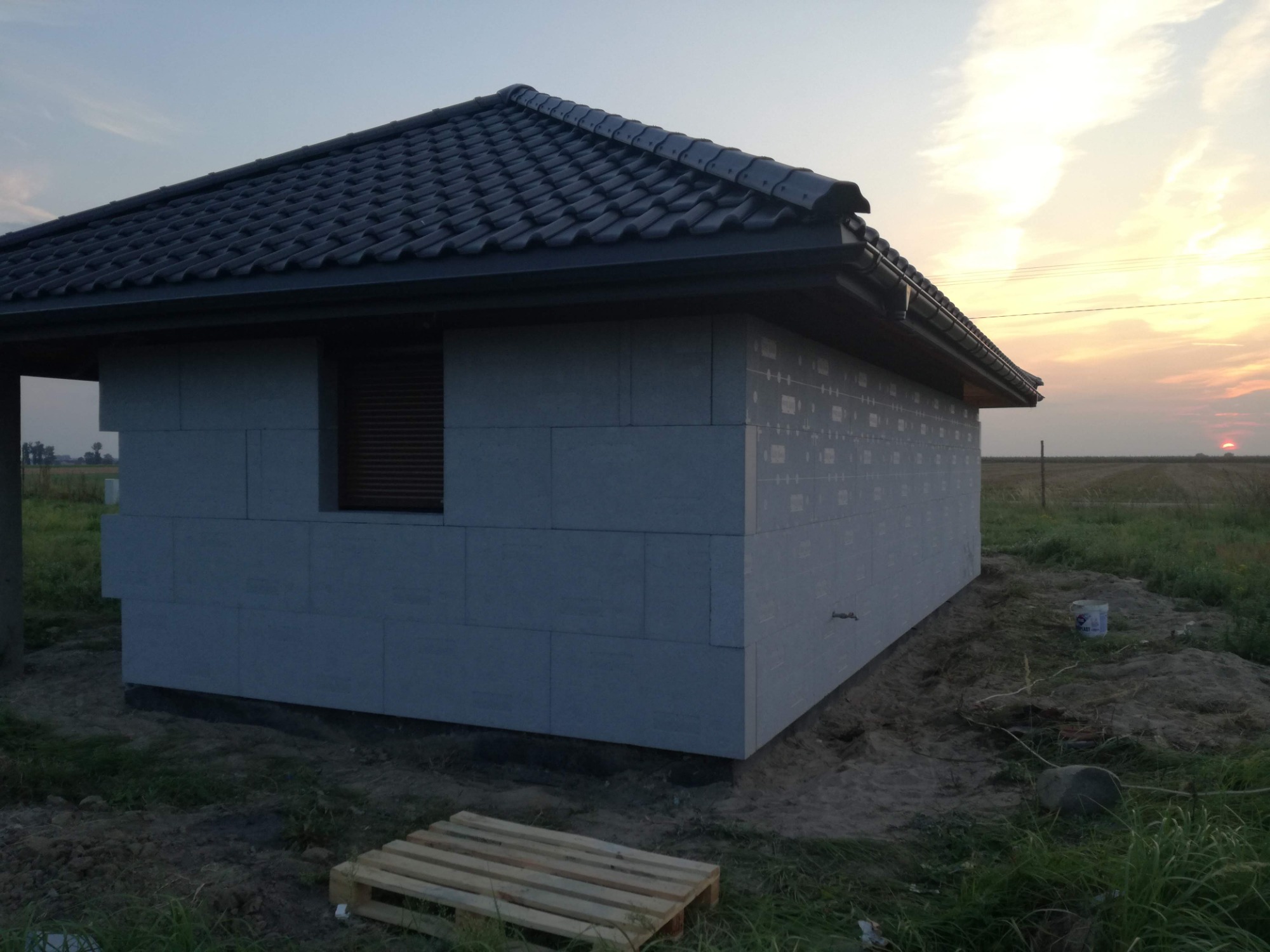 Niedokończony budynek z szarej cegły z oknem i ciemnym dachem z dachówki na tle zachodzącego słońca, widoczna paleta i wiadro na ziemi.