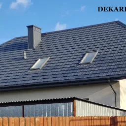 Dom jednorodzinny z ciemnoszarym dachem Ruukki z oknami dachowymi i kominem, widziany z perspektywy ogrodzenia. Widoczny fragment rusztowania.