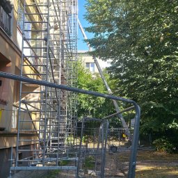 DEKARBUD ADAM MADZIAR - Budynek w rusztowaniach, zabezpieczony siatką. Widoczny podnośnik koszowy i zieleń. Słoneczny dzień, błękitne niebo.