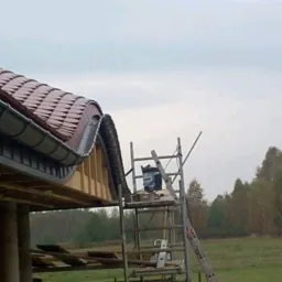 Montaż dachu z brązowej dachówki karpiówki i szarej rynny, widok z dołu na rusztowanie i częściowo wykończony budynek w tle. Praca w toku.