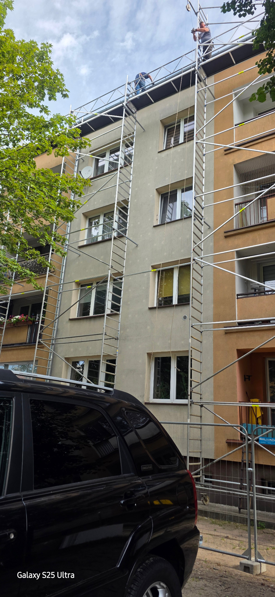 Praca na rusztowaniu przy elewacji budynku w Mińsku Mazowieckim. Dwóch mężczyzn na górze, widoczne okna i fragment zaparkowanego samochodu.