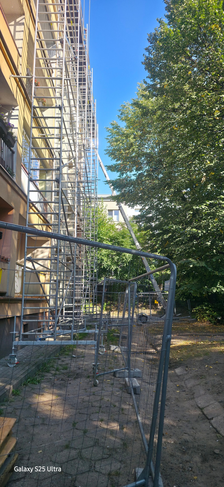 Budynek w rusztowaniach, zabezpieczony siatką. Widoczny podnośnik koszowy i zieleń. Słoneczny dzień, błękitne niebo.