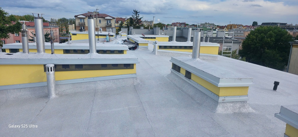 Szary dach płaski z żółtymi elementami i kominami wentylacyjnymi. Widok z góry na budynki w tle pod pochmurnym niebem.