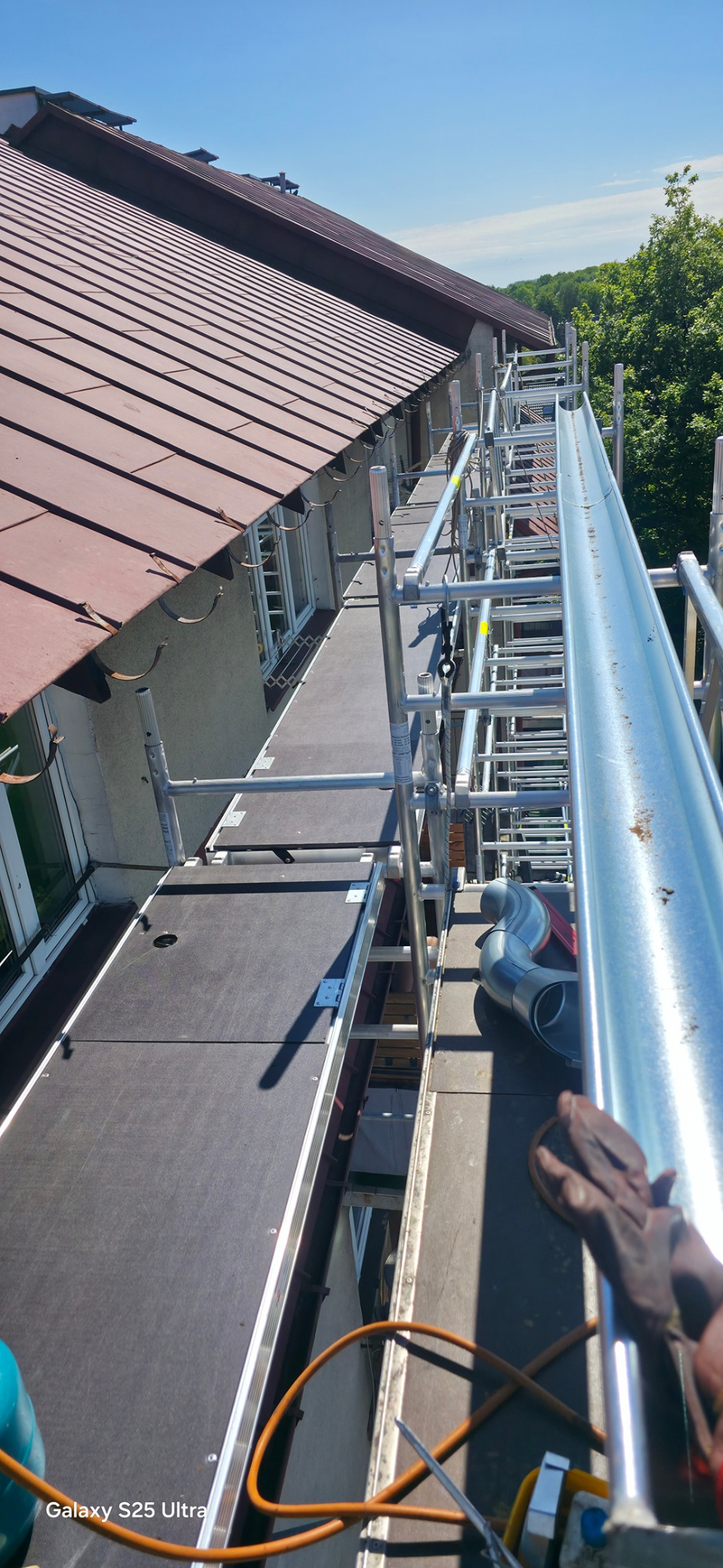 Montaż rynny na dachu domu z brązową dachówką, widok z rusztowania. Widoczne narzędzia i fragment rękawicy roboczej. Słoneczny dzień.