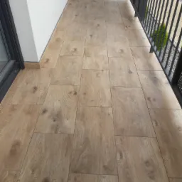 Wyłożony płytkami balkon z imitacją drewna, czarna metalowa balustrada, widok z góry na ułożone płytki.