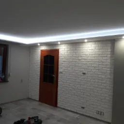 Pokój po remoncie z białą ceglaną ścianą, oświetleniem LED podsufitowym, drewnianymi drzwiami i szarymi ścianami; na podłodze narzędzia budowlane.