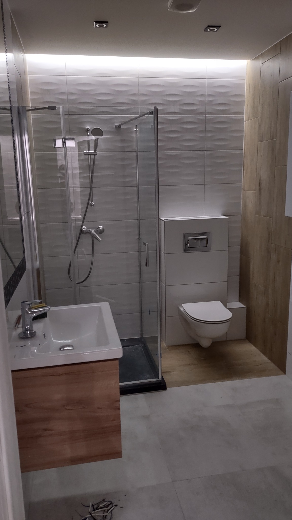 Nowoczesna łazienka po remoncie: kabina prysznicowa ze szklanymi drzwiami, umywalka z drewnianą szafką, podwieszana toaleta i płytki imitujące fale na ścianach.