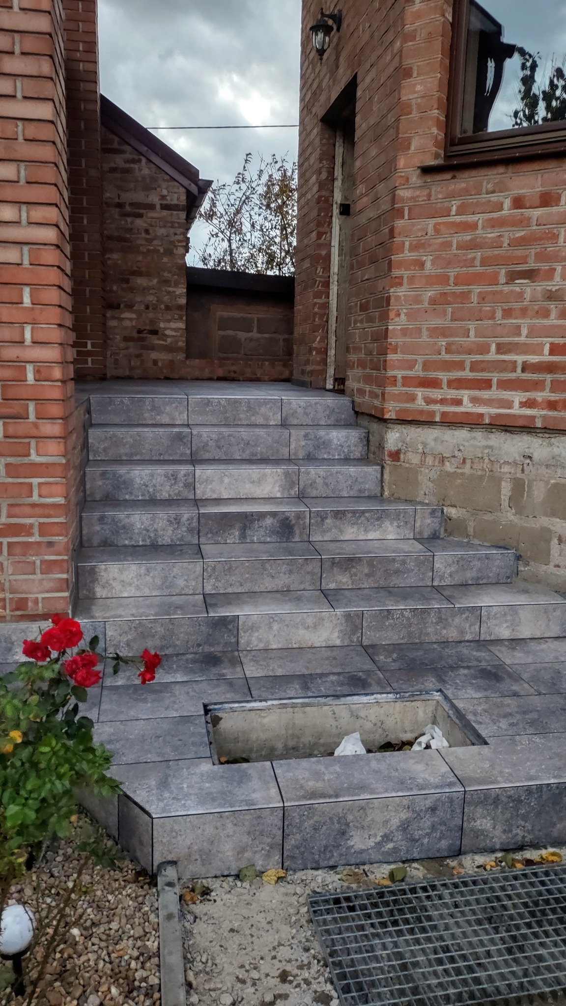 Wykończone płytkami schody wejściowe do domu z cegły, z widoczną kratką odpływową i czerwonymi różami po lewej stronie. Częściowo odkryty odpływ.