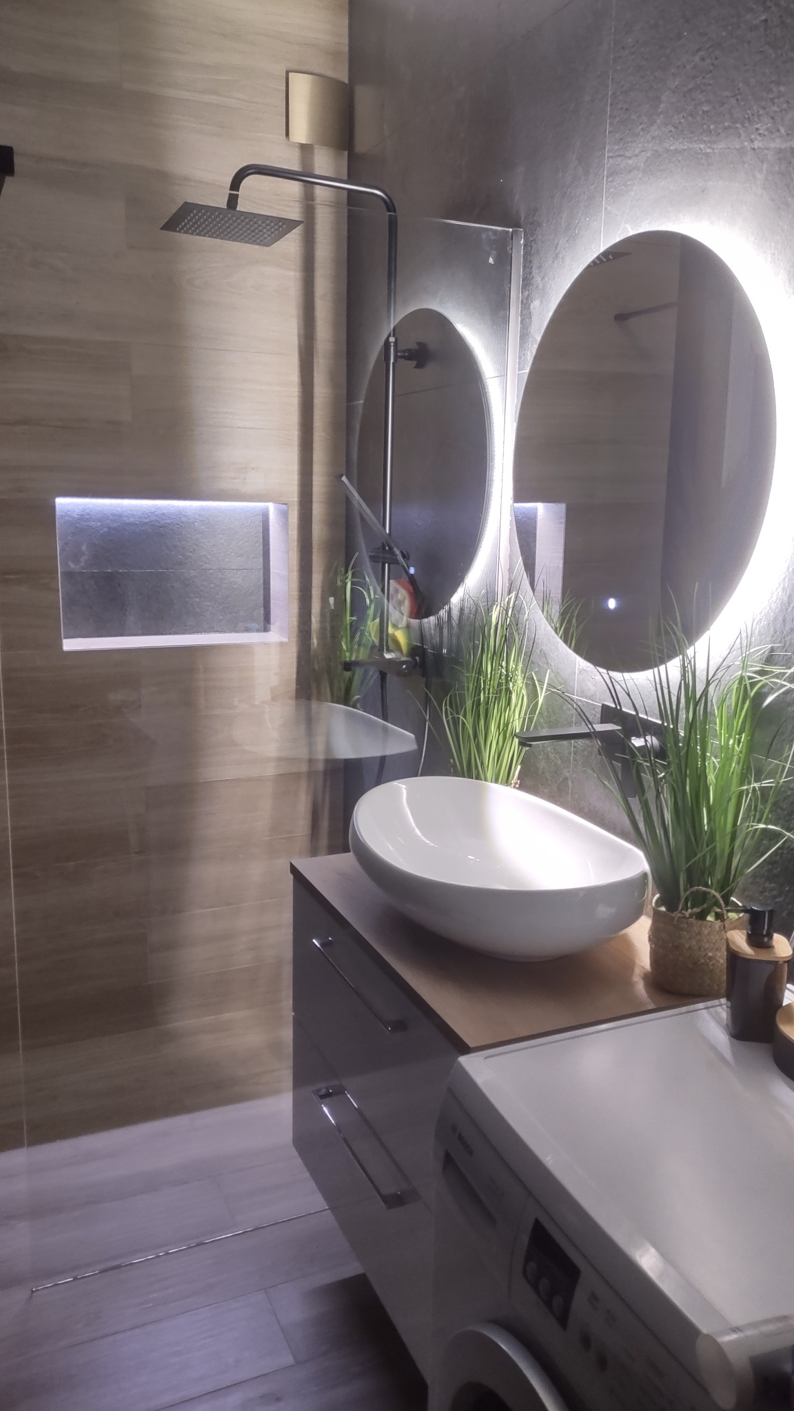 Nowoczesna łazienka z owalną umywalką, podświetlanym lustrem i deszczownicą. Minimalistyczny design z akcentami roślinnymi i szarą kolorystyką.