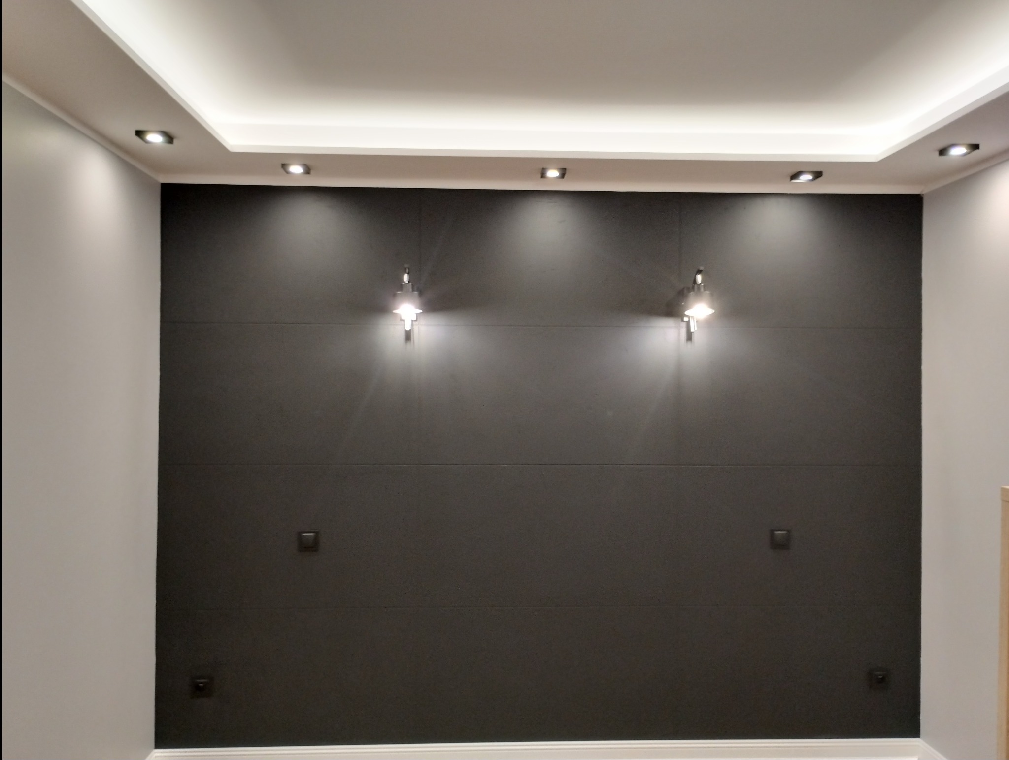 Nowoczesna ściana w kolorze grafitowym z subtelnym oświetleniem punktowym i dekoracyjnymi kinkietami. Minimalistyczny design z widocznymi włącznikami.
