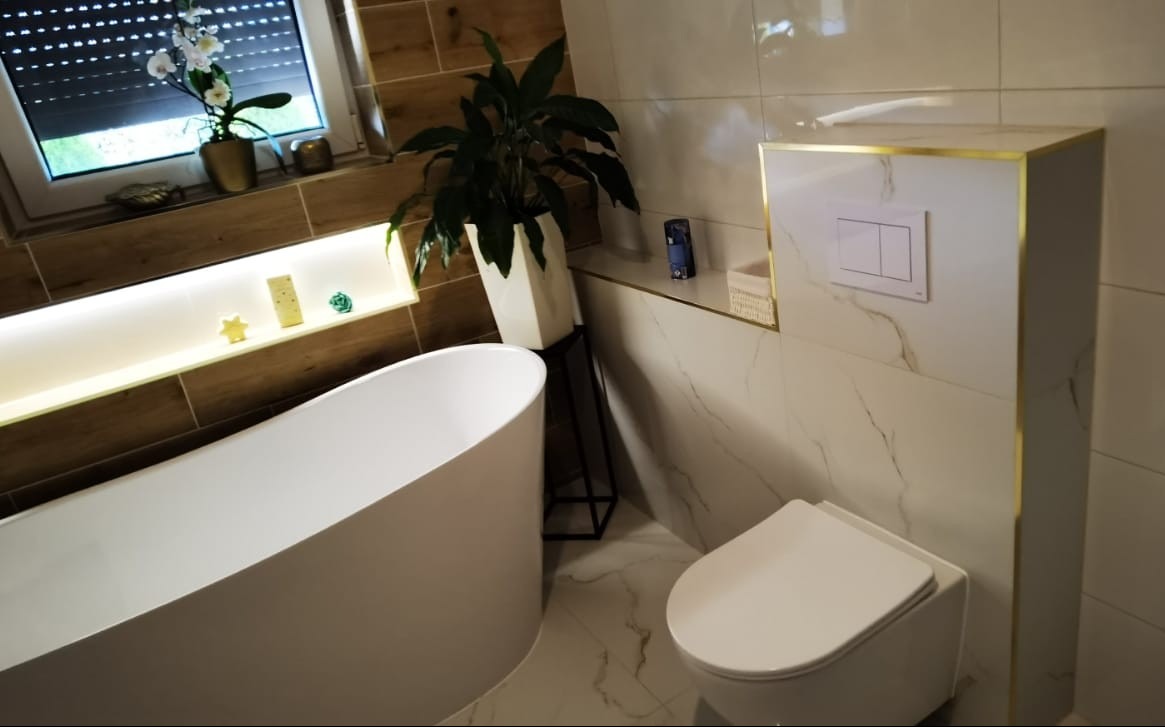 Nowoczesna łazienka z wolnostojącą wanną, marmurowymi płytkami i złotymi akcentami. Roślina doniczkowa dodaje świeżości wnętrzu.